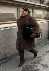 Brun pälsjacka, långa ärmar, svart handväska, bruna leggings, knähöga stövlar. Kvinna som går mot en texturerad, ljust färgad vägg.