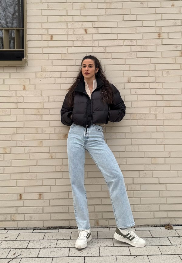 Giacca puffer nera cropped con colletto alto, abbinata a jeans alla caviglia blu chiaro e sneakers bianche con dettagli verdi.