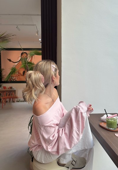 Sudadera rosa claro sin hombros, falda blanca con patrón floral, calcetas de tobillo gris claro y zuecos beige, con una bebida sobre una mesa.