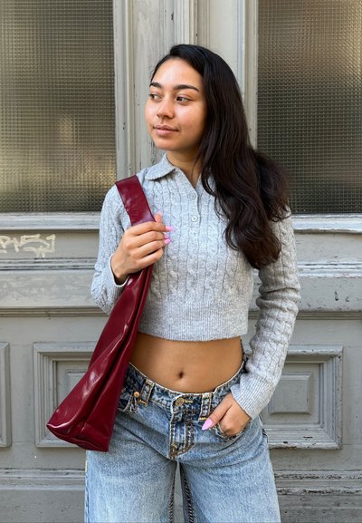 Suéter cropped de punto trenzado gris, jeans de tiro alto azules y un bolso burdeos brillante. Textura suave con detalles de botones y diseño ajustado.