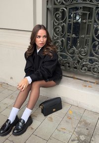 Chaqueta negra oversized, camisa blanca con cuello, falda negra, medias transparentes y mocasines negros con un acabado texturizado. Bolso negro al lado.