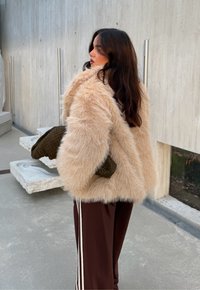 Cappotto in pelliccia sintetica color beige chiaro con una consistenza spessa, maniche lunghe e vestibilità ampia. Abbinato a pantaloni marroni con strisce bianche laterali e una borsa verde.