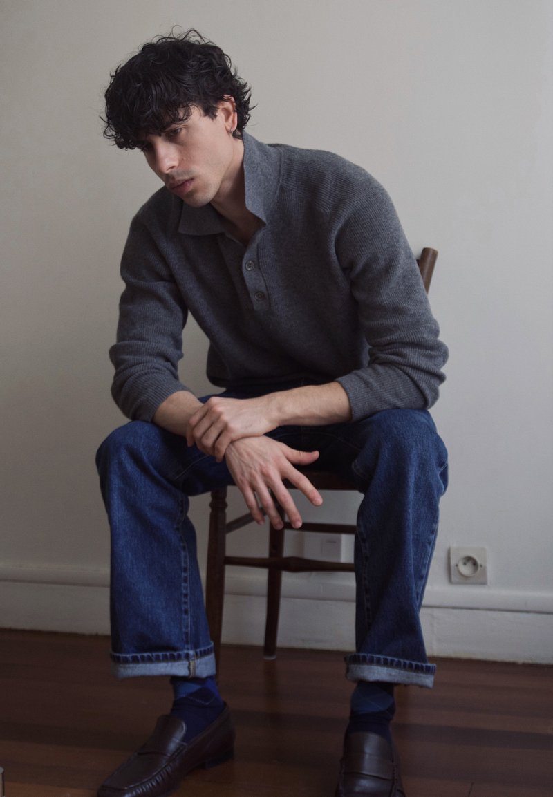 Young man with curly hair, wearing a gray sweater and rolled-up jeans, sitting on a wooden chair with hands clasped, looking down.