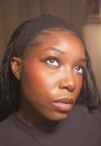 Nahaufnahme eines Gesichts mit dunkler Haut, das ein schwarzes Stirnband trägt. Zu den Merkmalen gehören lange Wimpern, ein subtiler Schimmer auf der Haut und ein leicht rosa Rouge.