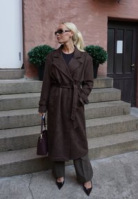 Smeđa suedna trench kaput s remenom, dvostruko kopčanje, široki reveri, uparen s tamnim hlačama i burgundi torbicom.