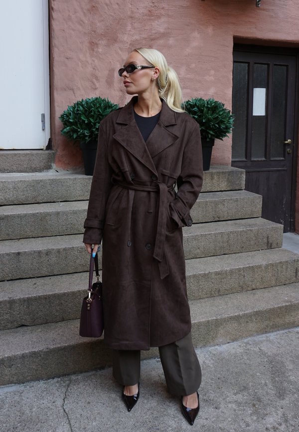 Brauner Wildleder-Trenchcoat mit Gürtel, doppelreihigem Design, breiten Revers, kombiniert mit dunklen Hosen und einer bordeauxfarbenen Handtasche.