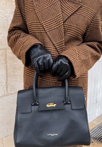 Personne portant des gants en cuir noir et un manteau à carreaux marron tenant un sac à main noir Lancaster Paris par ses poignées.