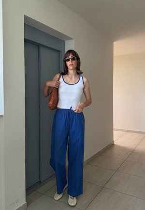 Mujer con gafas de sol, camiseta sin mangas blanca, pantalones azules, sosteniendo un bolso marrón tejido, de pie en el interior cerca de puertas grises cerradas de ascensor.