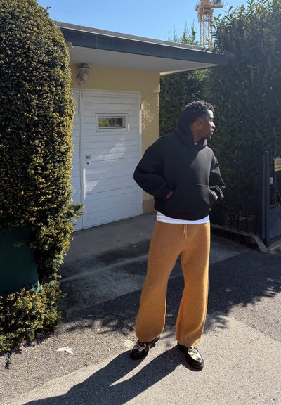 Persona con sudadera negra con capucha y pantalones marrones de pie afuera cerca de arbustos podados y una puerta blanca, mirando hacia la derecha bajo la luz del sol.