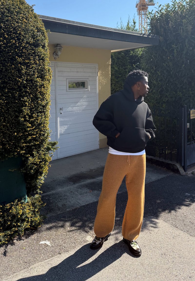 Personne en sweat à capuche noir et pantalon marron debout à l'extérieur près de buissons taillés et d'une porte blanche, regardant vers la droite au soleil.