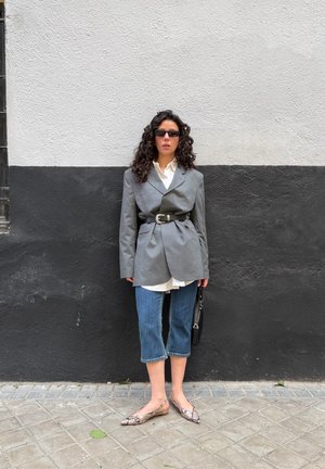 Mujer con cabello rizado, lleva gafas de sol, blazer gris oversize ceñido a la cintura, camisa blanca, vaqueros crop, zapatos planos de piel de serpiente, y sostiene un bolso negro.