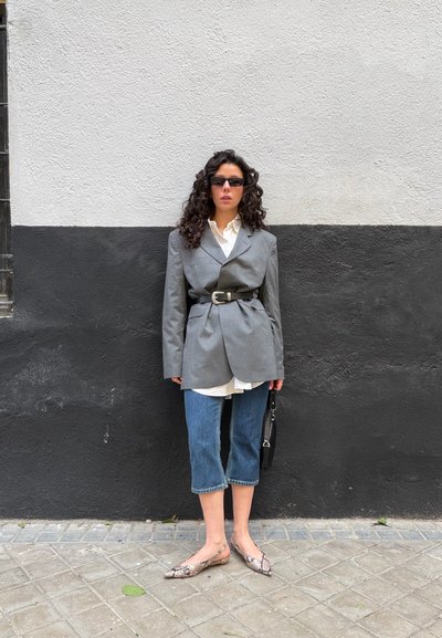 Mujer con cabello rizado, lleva gafas de sol, blazer gris oversize ceñido a la cintura, camisa blanca, vaqueros crop, zapatos planos de piel de serpiente, y sostiene un bolso negro.