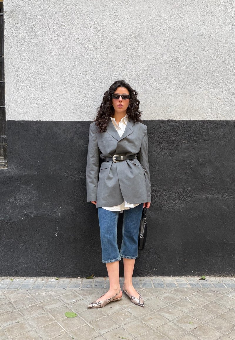 Mujer con cabello rizado, lleva gafas de sol, blazer gris oversize ceñido a la cintura, camisa blanca, vaqueros crop, zapatos planos de piel de serpiente, y sostiene un bolso negro.