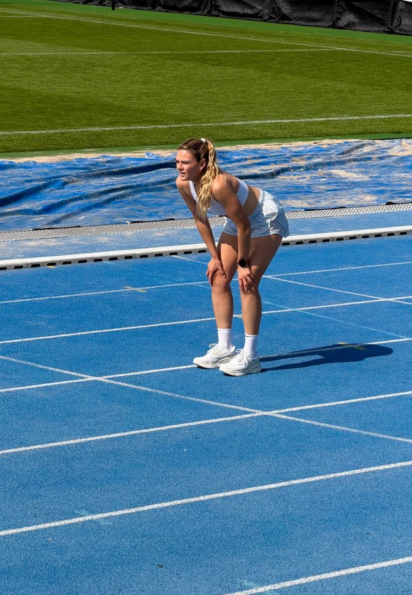 Athlète en tenue de sport blanche penché en avant, mains sur les genoux, debout sur une piste bleue près d'un terrain d'herbe verte.