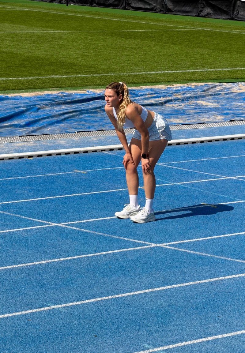 Спортист в бяло спортно облекло, наведени напред с ръце на коленете, стои на синя писта до зелено тревно поле.