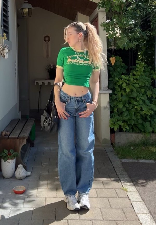 Roheline lühikesed Adidas t-särk, kõrge taljega heledad sinised teksad, valged tennisekingad, must kott. Väliskeskkond roheluse ja kivist teega.