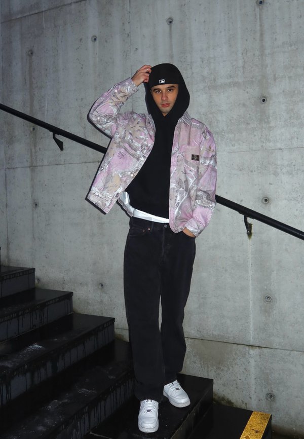 Casaco rosa claro e estampado por cima de um hoodie preto, combinado com jeans pretos e ténis brancos, de pé em escadas escuras contra uma parede de betão.