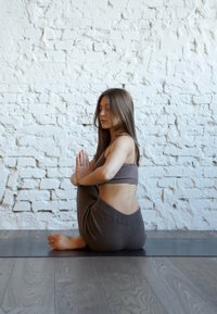 Szürke sportöltözéket viselő nő keresztbe tett lábbal ül egy jógaszőnyegen, összetett kézzel, csukott szemmel meditál egy fehér téglafal előtt.