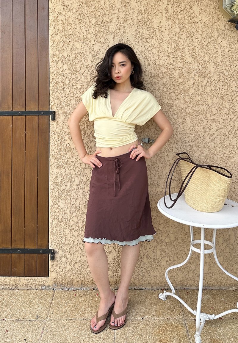Femme aux cheveux foncés portant un haut court jaune clair et une jupe marron se tient les mains sur les hanches à côté d'une table ronde blanche avec un sac en paille.