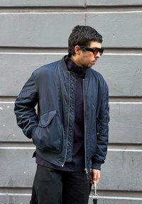 Άνδρας που φοράει μαύρο μπουφάν bomber και γυαλιά ηλίου στέκεται μπροστά σε γκρίζο τοίχο, κρατώντας με το δεξί χέρι έναν ιμάντα καρπού με μεταλλικό δαχτυλίδι.