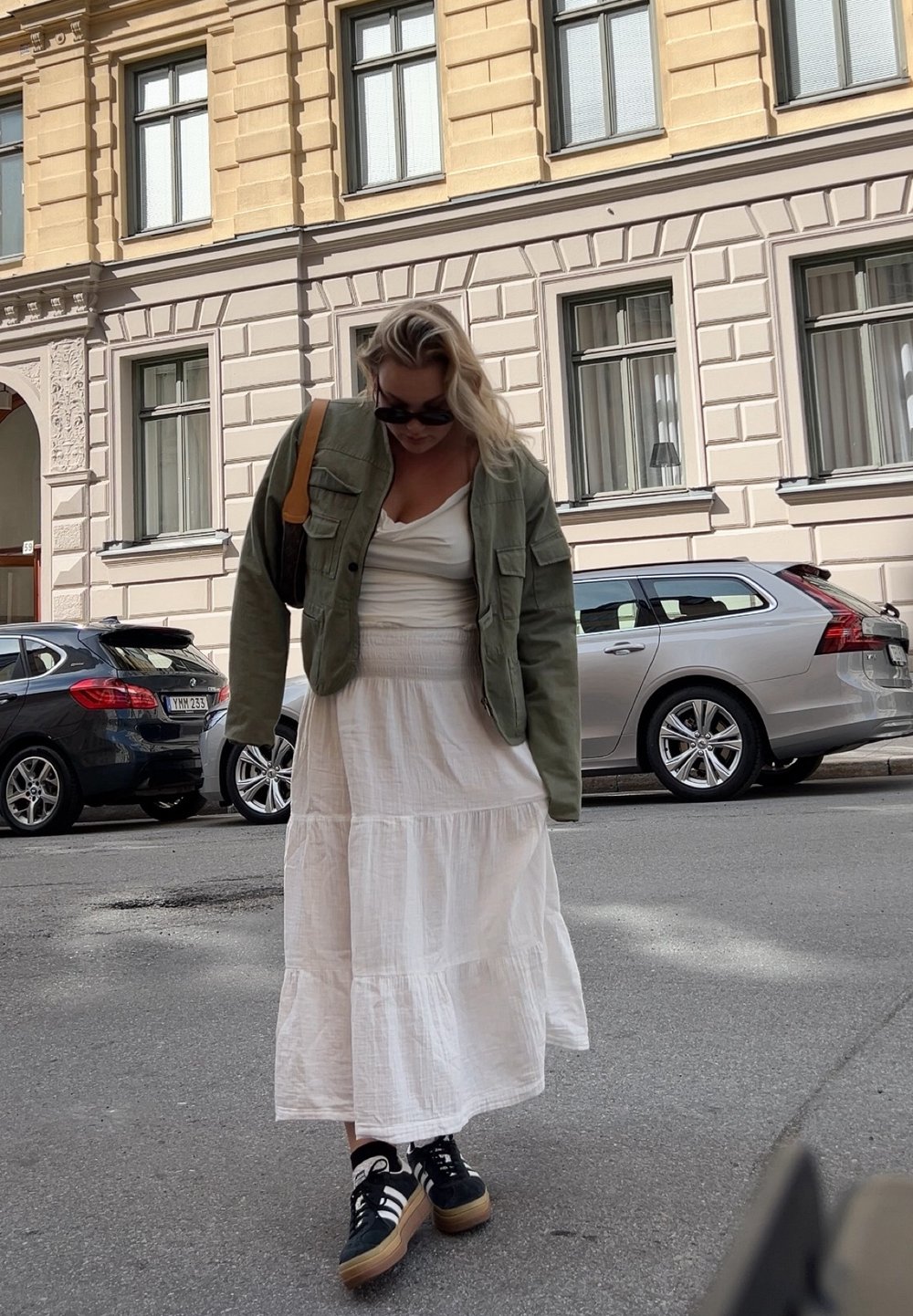 Olivově zelená vojenského stylu bunda, bílá vrstvená šaty a černé tenisky s hnědými akcenty. V pozadí jsou zaparkovaná auta a fasáda budovy.
