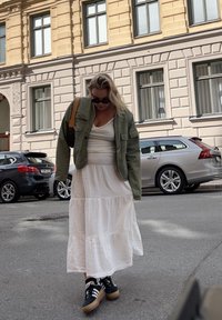 Olivově zelená vojenského stylu bunda, bílá vrstvená šaty a černé tenisky s hnědými akcenty. V pozadí jsou zaparkovaná auta a fasáda budovy.