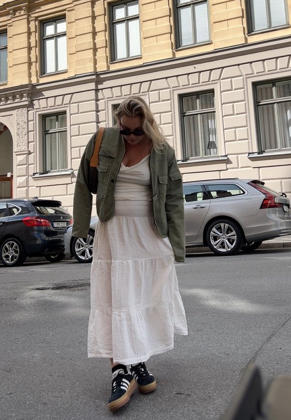 Olivovo zelená vojenská bunda, biela šaty v tichých vrstvách a čierne tenisky s hnedými akcentmi. Pozadie zobrazuje zaparkované autá a fasádu budovy.