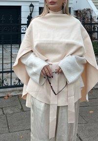 Svijetlo ružičasti vuneni pončo sa širokim rukavima i remenom u struku, nošen preko sjajne srebrne suknje. Naočale lagano držane u rukama.