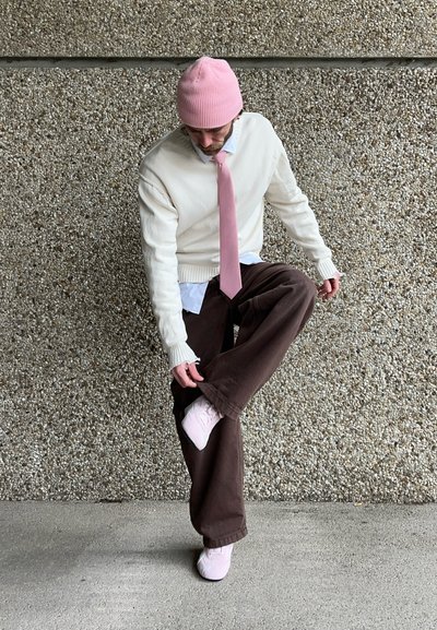 Suéter de punto off-white, corbata rosa pastel, pantalones anchos marrones y zapatillas rosa claro. Los accesorios incluyen un gorro rosa.