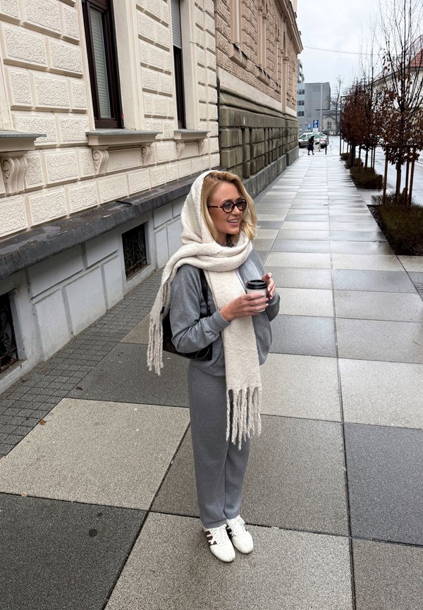 Frau in grauem Outfit und großem weißem Schal, die eine Kaffeetasse hält und auf einem gefliesten Bürgersteig in der Stadt nahe eines beigen Gebäudes steht.