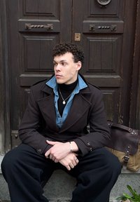 Jeune homme aux cheveux bouclés, portant plusieurs vestes superposées et des bijoux, assis sur une marche en pierre devant de grandes portes en bois sombre, à côté d'un sac.