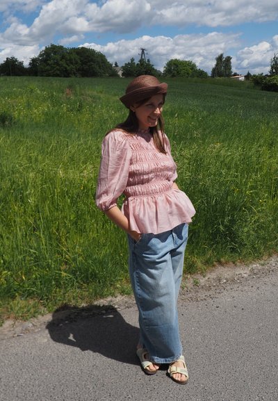 Mujer con un sombrero marrón, blusa rosa y jeans azules anchos, de pie en una carretera junto a un campo de hierba verde bajo un cielo parcialmente nublado.