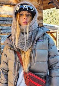 Puffy grijze jas met rits, dikke grijze sjaal, zwarte pet met logo en rode schoudertas. Soepele textuur, warme materialen, gelaagde uitstraling.