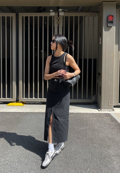 Top negro sin mangas combinado con una larga falda de mezclilla negra con una abertura en el frente. Los accesorios incluyen un pequeño bolso negro de hombro y zapatillas deportivas.