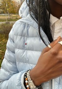 Šviesiai mėlynas pūkinis švarkas su mažomis raudonomis, baltomis ir mėlynomis logotipų. Sidabriniai aksesuarai ir tekstūruotas laikrodis ant riešo su apyrankėmis.