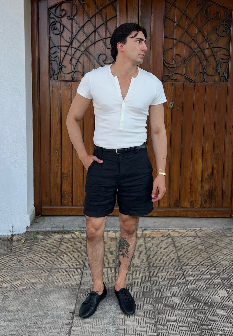 Jeune homme regardant à droite, portant une chemise blanche à manches courtes, un short noir, des chaussures noires, avec un tatouage sur le mollet gauche, debout sur un sol carrelé.