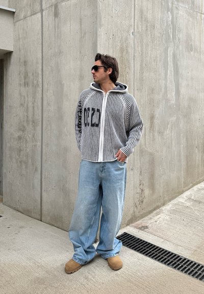 Sudadera con cremallera a rayas y capucha, que presenta un gráfico en la manga izquierda, combinada con jeans de pierna ancha de color azul claro y zuecos color miel.