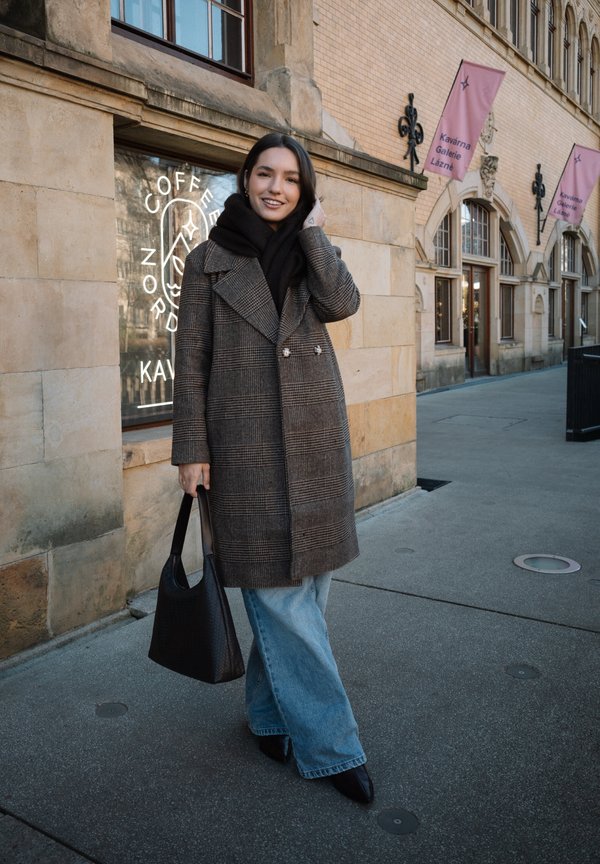 Donna sorridente, in piedi fuori da un caffè, indossa un cappotto a quadri, una sciarpa nera, jeans blu, scarpe nere e tiene una borsa nera.