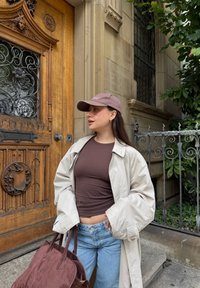 Beige trenchcoat over en brun tætsiddende top, lyseblå jeans og en brun ruskindstaske. Blød tekstur og neutrale farver med en kasket og tilbehør.