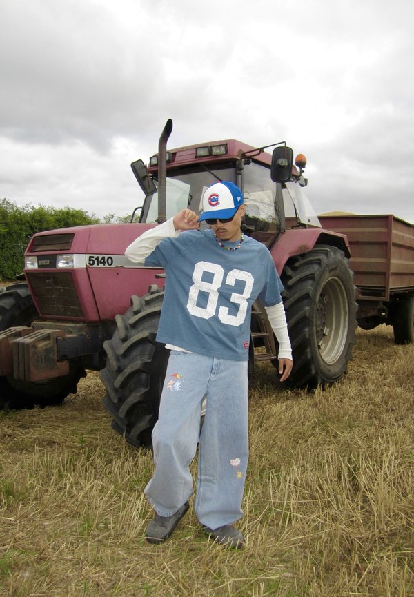Person iført en blå kasket, en stor skjorte og jeans, stående foran en rød traktor på en mark under en overskyet himmel.