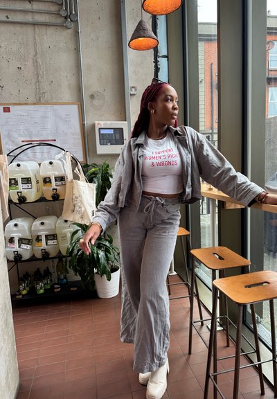 Chaqueta de lino gris y pantalones a juego, camisa blanca con texto rojo y zapatos de plataforma blancos. Entorno interior con plantas y acentos de madera.