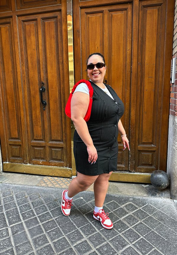 Vestido preto risca de giz com botões, combinado com uma t-shirt branca e uma bolsa vermelha. Usa óculos de sol e ténis vermelho e branco. De pé sobre calçada.