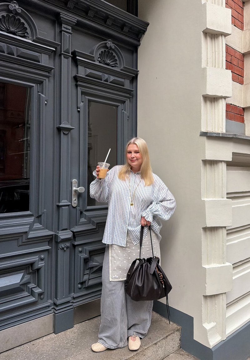 Femme blonde debout près d'une porte ornée gris foncé, portant un chemisier rayé, un pantalon large gris, tenant un café glacé et un sac à main noir.