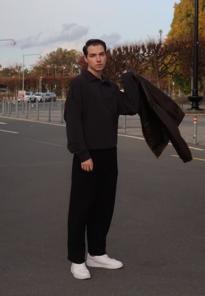 Camisa oscura de manga larga con cuello, pantalones negros holgados y zapatillas blancas. La persona sostiene una chaqueta marrón en la mano izquierda.