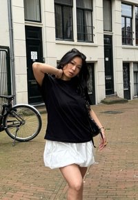 T-shirt noir en coton associé à une mini-jupe blanche texturée. Les accessoires comprennent un petit sac noir. L'arrière-plan présente un pavé en brique et un vélo.