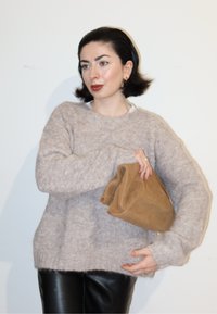 Femme aux cheveux courts et foncés portant un pull beige et un pantalon noir, tenant une pochette souple marron sur un fond blanc.
