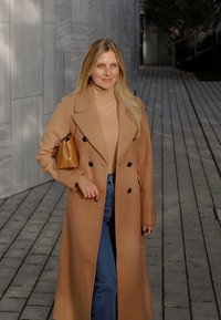 Kamel-farbener Wollmantel mit doppelter Knopfleiste, kombiniert mit einem beigen Rollkragenpullover und blauen Jeans. Trägt eine kleine braune Handtasche.