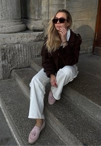 Kvinde med langt blond hår iført store solbriller, mørk frakke, hvide bukser og lyserøde frynse-loafers, der sidder på stenskader ved en tekstureret væg.