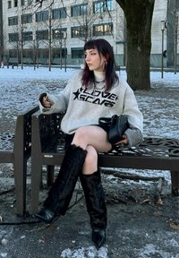 Suéter cinza oversized com o texto "LOVE SCARS", perneiras de pelúcia negra, botas pretas de bico fino e uma mala preta, sentada num banco de parque.