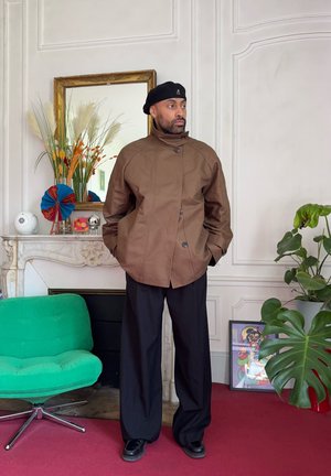 Hombre con chaqueta marrón, boina negra y pantalones negros anchos está de pie dentro de una habitación cerca de una silla verde, chimenea, plantas y una obra de arte enmarcada sobre una alfombra roja.